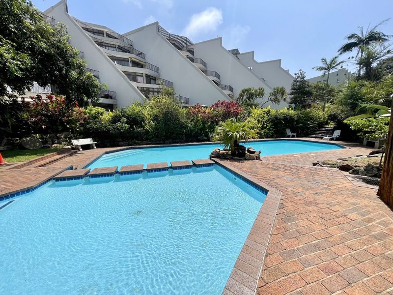 A well-maintained outdoor swimming pool area featuring brick paving and lush landscaped gardens, set against the backdrop of a multi-story apartment complex.