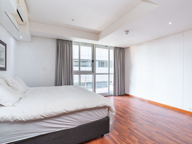 A bright and spacious bedroom featuring warm laminate flooring, recessed ceiling lighting, a wall-mounted air conditioner, and large sliding glass doors leading to a balcony.