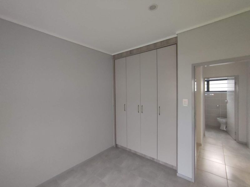 A clean, modern bedroom featuring neutral grey walls, tiled flooring, and ample built-in cupboard storage, with an adjacent view into a tiled bathroom.