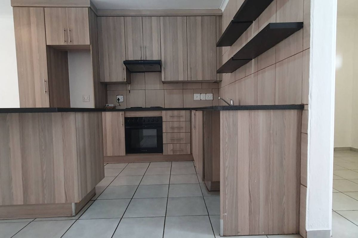 A clean, modern kitchen featuring wood-look cabinetry, dark countertops, built-in oven with extractor fan, and tiled flooring in an open-plan layout.