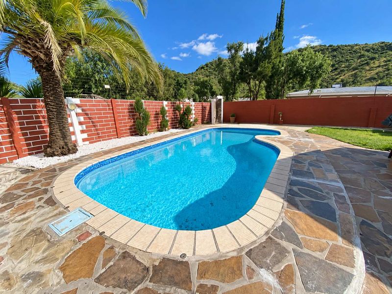 A well-maintained outdoor swimming pool area featuring stone paving, a brick boundary wall, and landscaped surroundings with a palm tree.