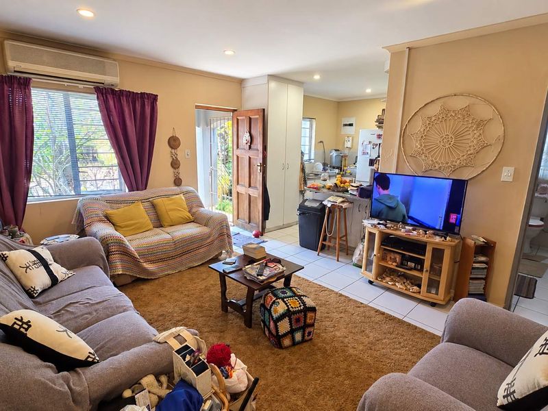 Open-plan living area featuring tiled flooring, recessed downlighting, and a wall-mounted air conditioning unit, with direct access to the kitchen and adjacent bathroom.