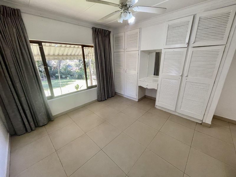A bright, empty bedroom featuring built-in louvered cupboards, a vanity station, ceiling fan, and large windows overlooking the garden.