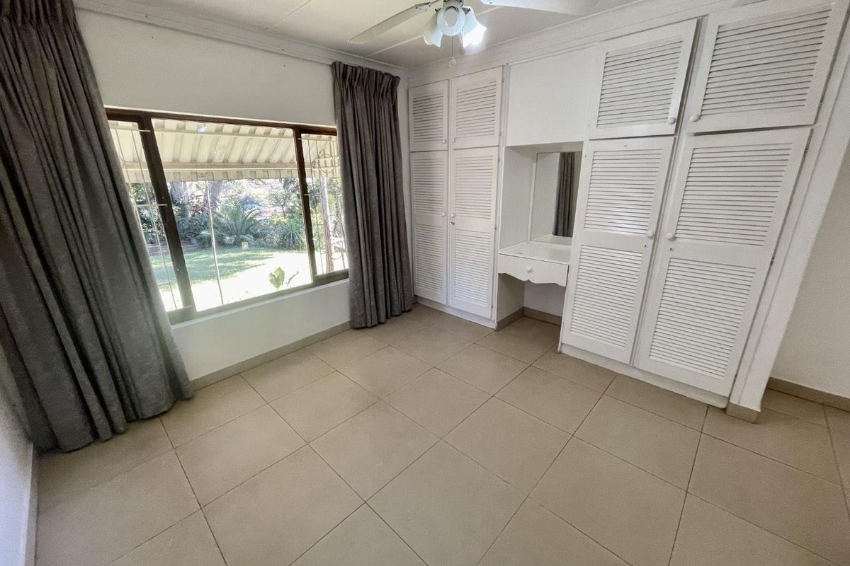 A bright, empty bedroom featuring built-in louvered cupboards, a vanity station, ceiling fan, and large windows overlooking the garden.