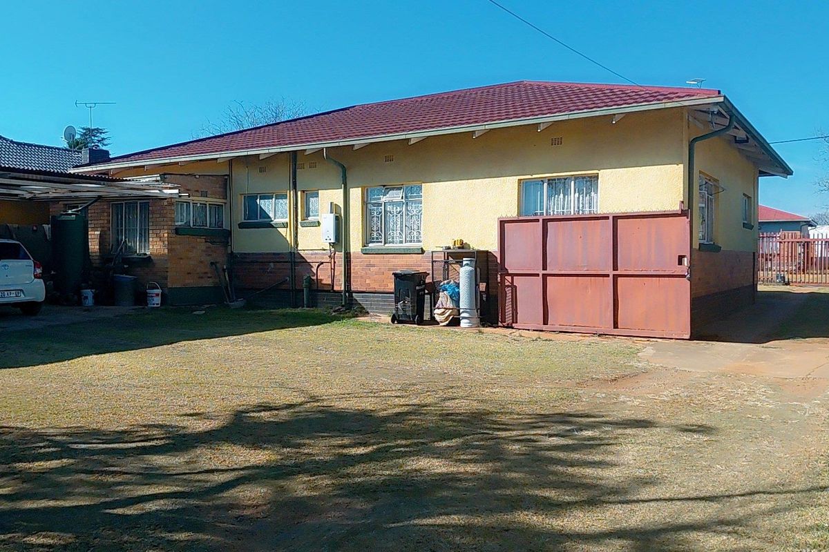 Rear view of a single-story house featuring a metal roof, brick and plaster facade, attached carport, and a large backyard area.