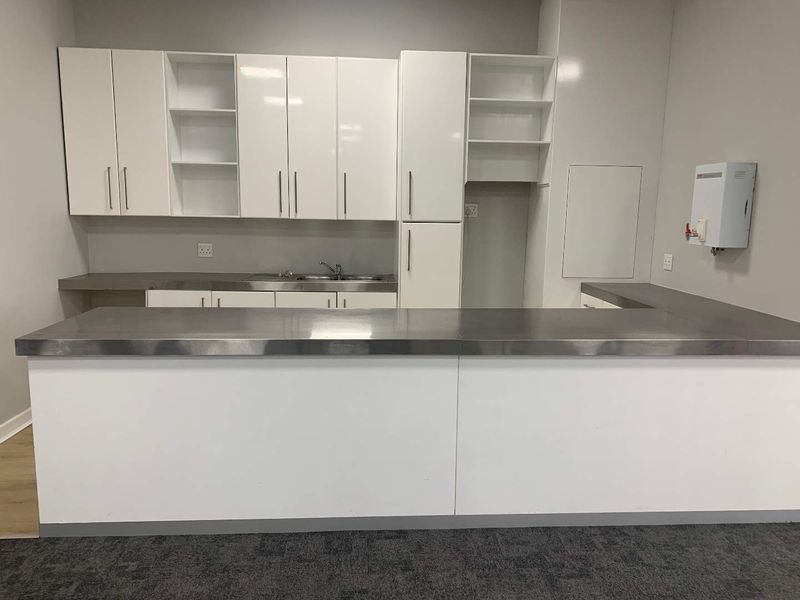 A clean, modern office kitchenette featuring white cabinetry, stainless steel countertops, a sink, and a wall-mounted water heater.