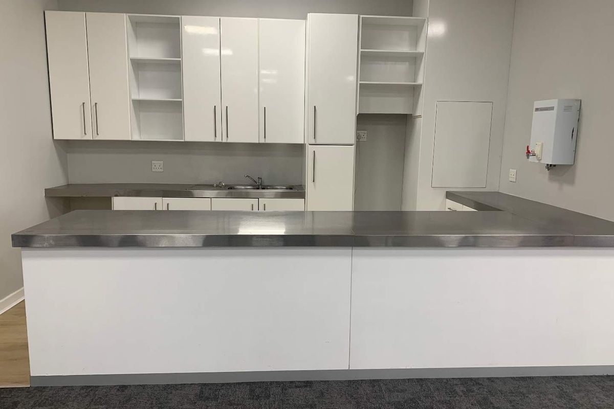 A clean, modern office kitchenette featuring white cabinetry, stainless steel countertops, a sink, and a wall-mounted water heater.