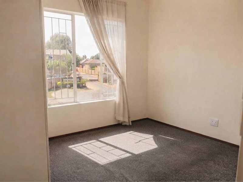 A bright, empty bedroom featuring carpeted flooring, a large window with security bars, and neutral wall finishes.