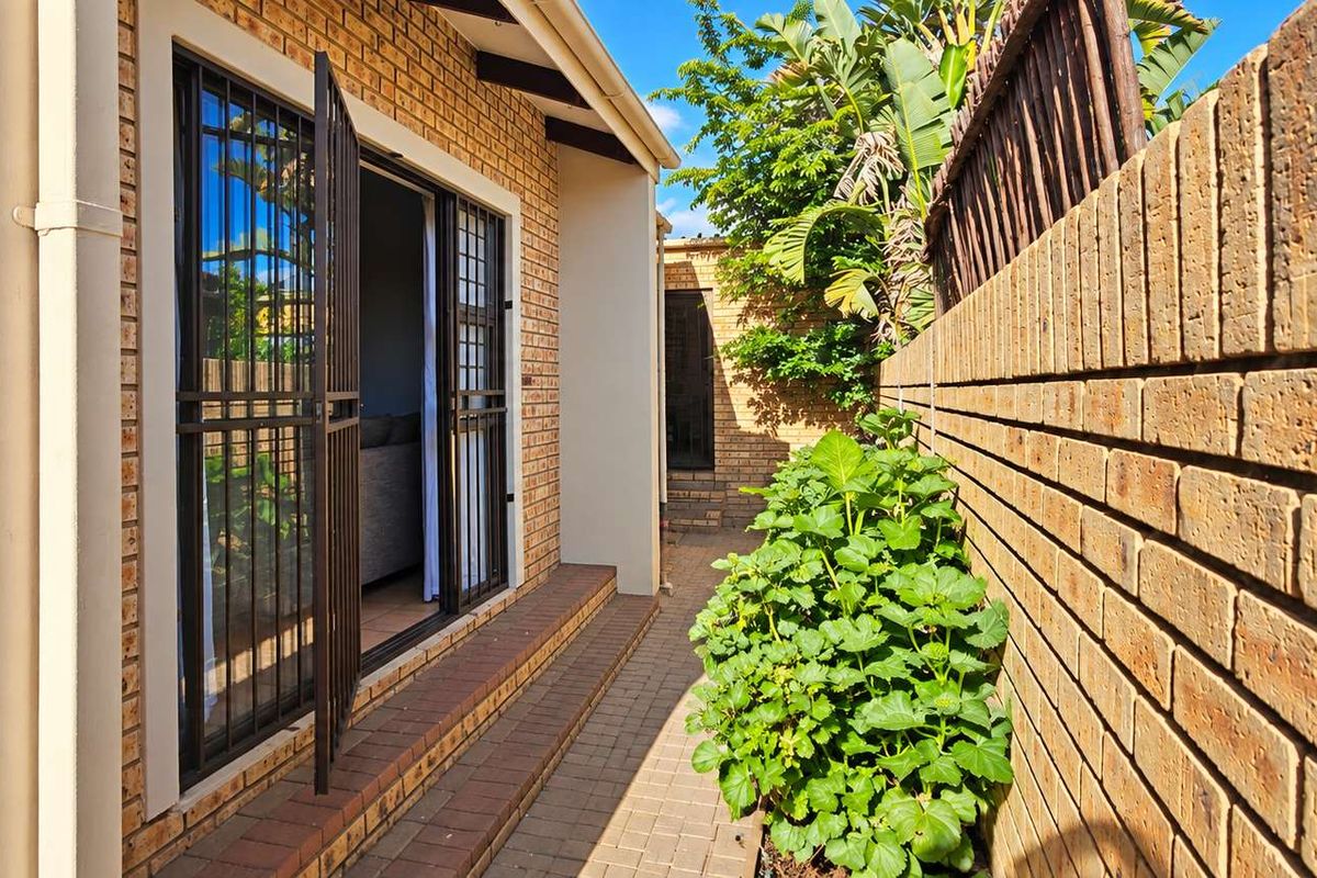 A side exterior view of the townhouse featuring a paved walkway, brick boundary wall, and a secure sliding door entrance.