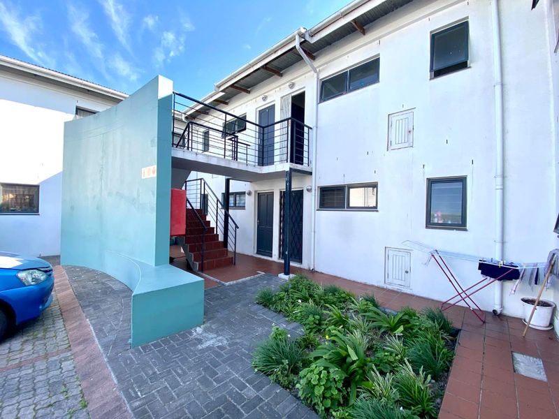 Rear exterior view of the apartment building featuring a paved courtyard, garden bed, and external staircase leading to the upper level balcony.