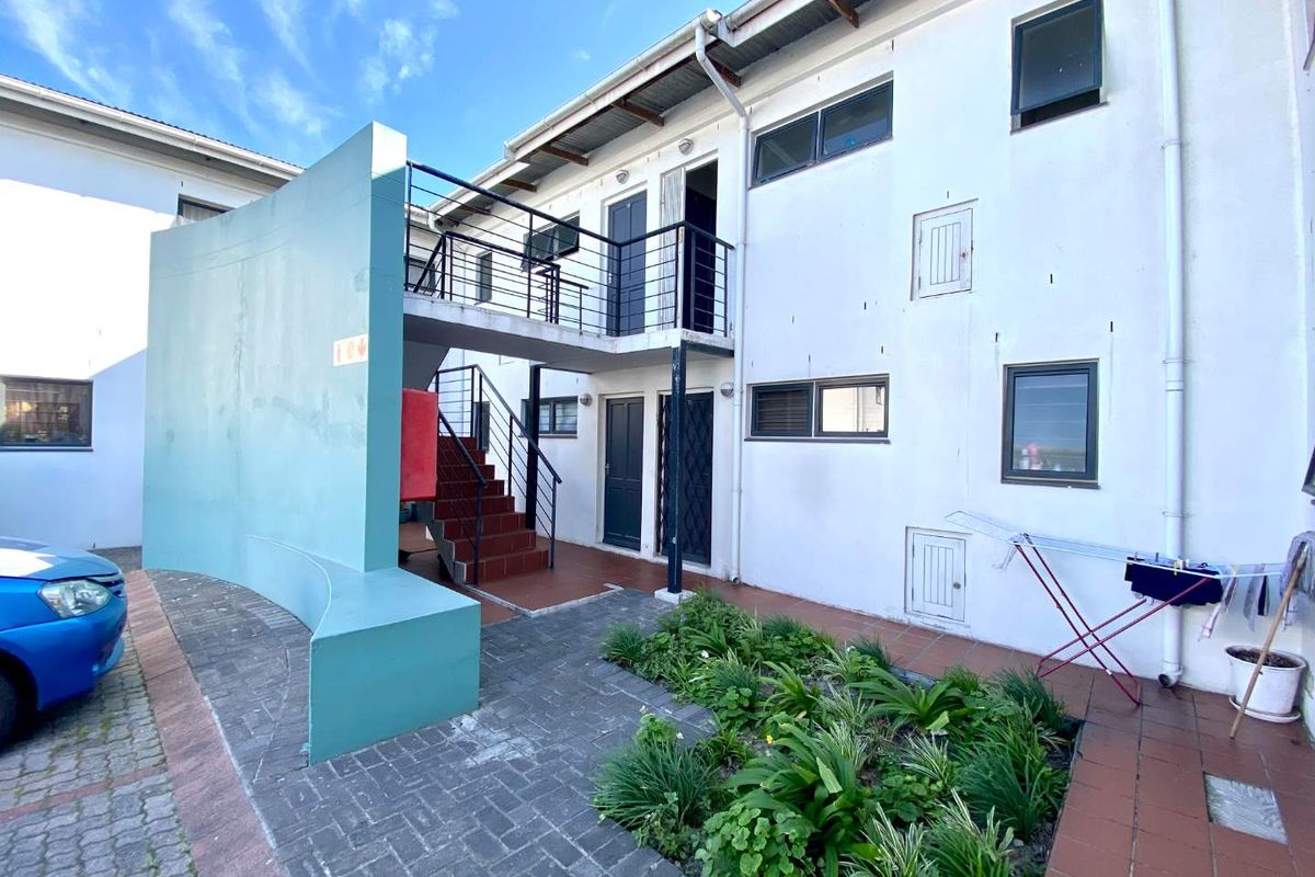 Rear exterior view of the apartment building featuring a paved courtyard, garden bed, and external staircase leading to the upper level balcony.