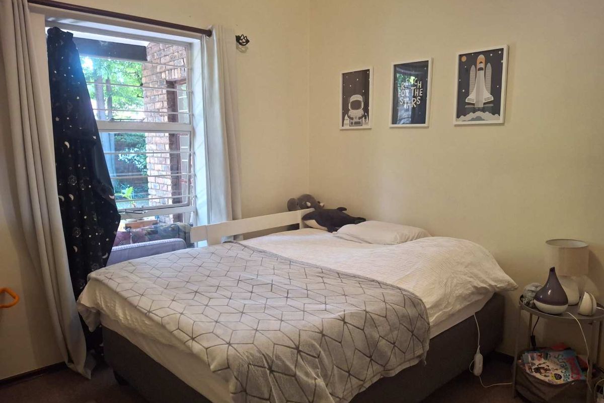 A functional bedroom featuring a large window with security bars, providing natural light and a view of the exterior brickwork.