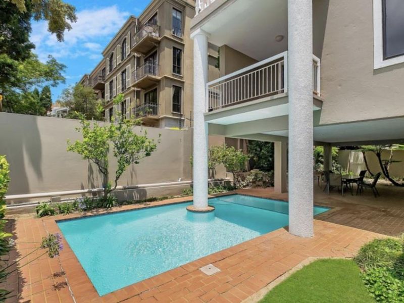Outdoor pool area featuring a swimming pool, brick paving, and a covered patio space beneath an elevated balcony structure.
