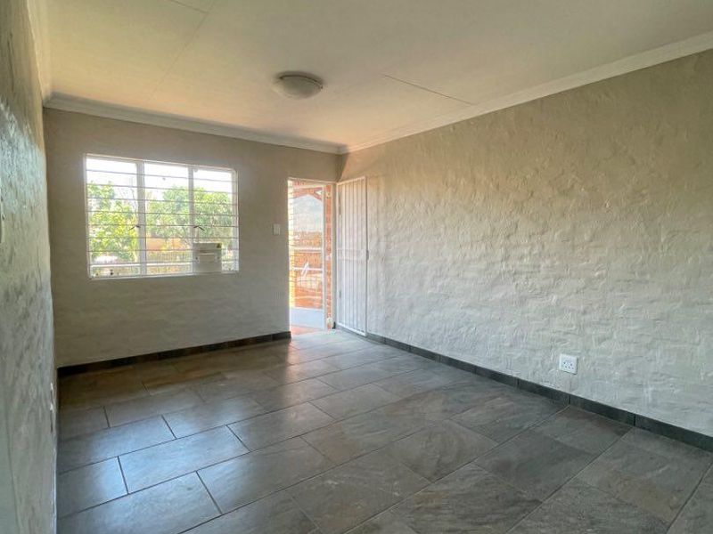 An empty living room featuring neutral textured brick walls and large format floor tiles, with natural light entering through a window and exterior door.