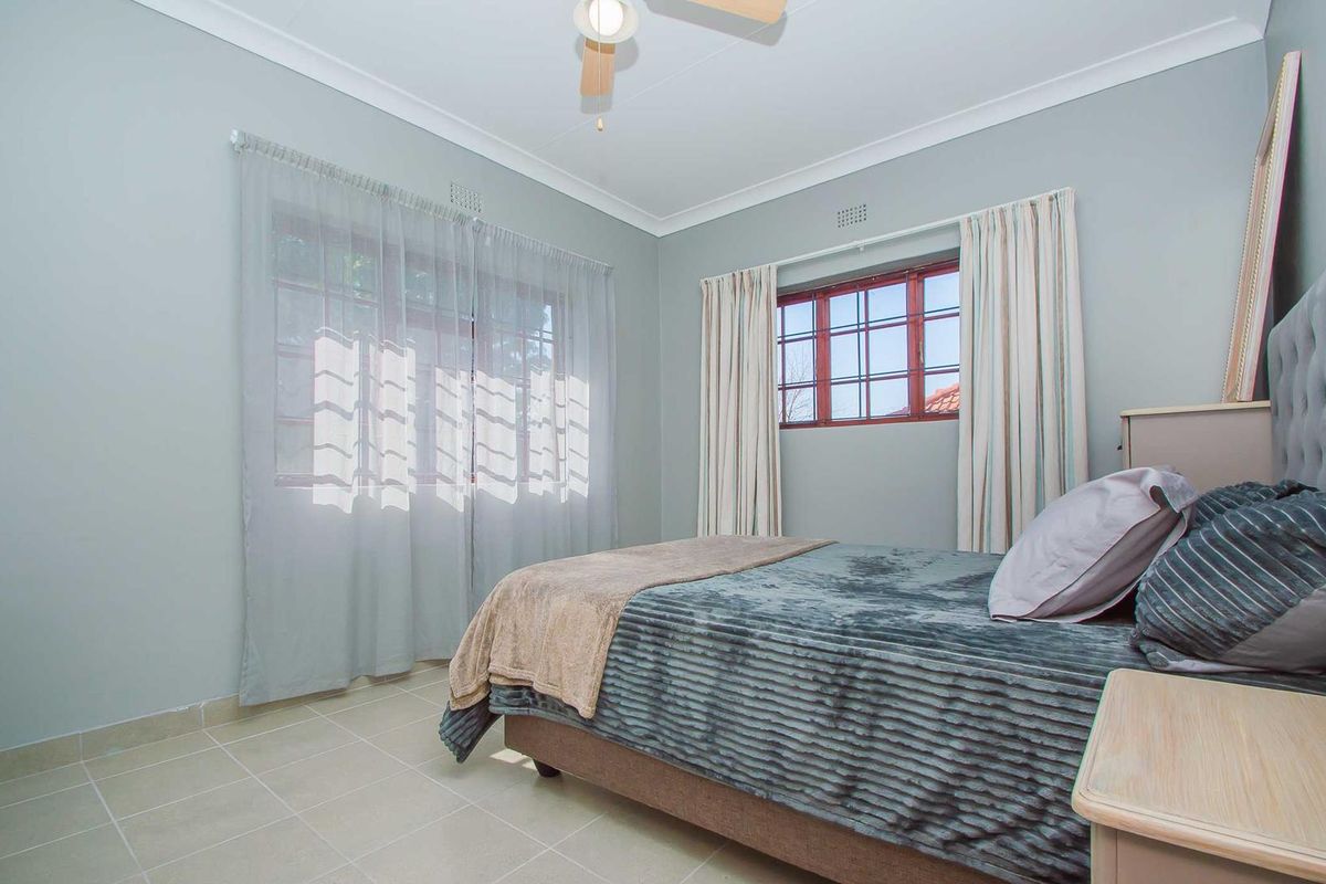 A well-maintained bedroom featuring tiled flooring, neutral wall tones, a ceiling fan, and large windows with fitted curtain tracks.