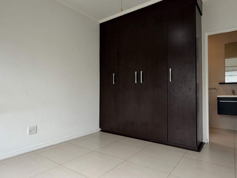 A clean, empty bedroom featuring dark wood built-in wardrobes, neutral tiled flooring, and direct access to an en-suite bathroom.