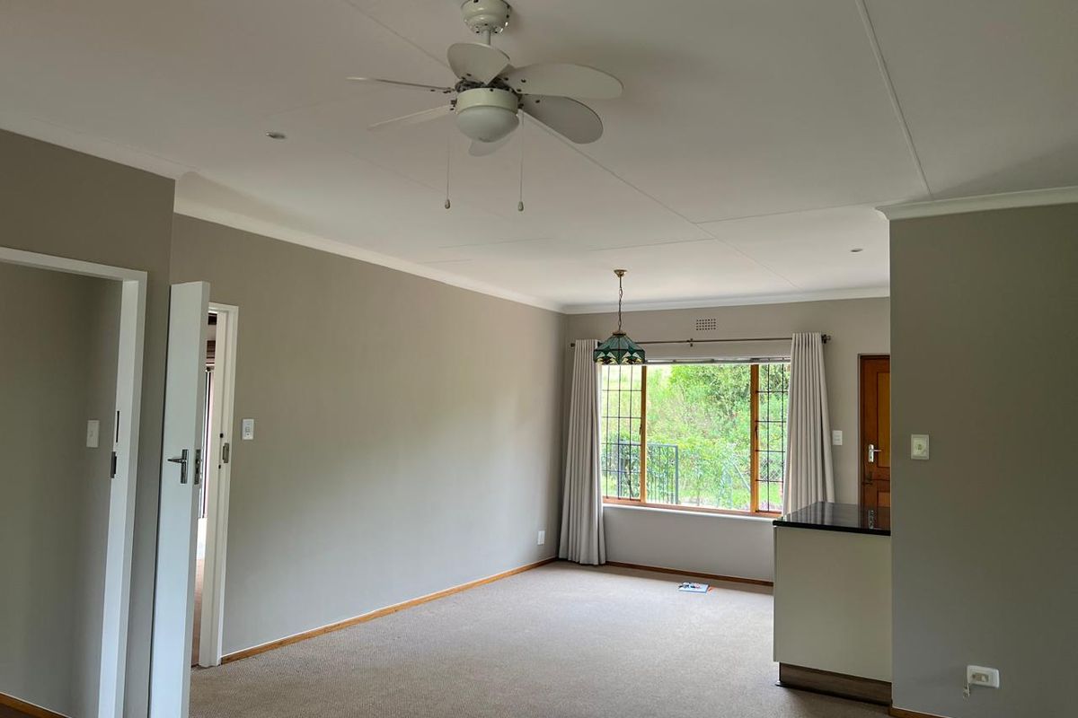 A spacious, empty living area featuring neutral wall tones, carpeted flooring, a ceiling fan, and large windows providing natural light and garden views.