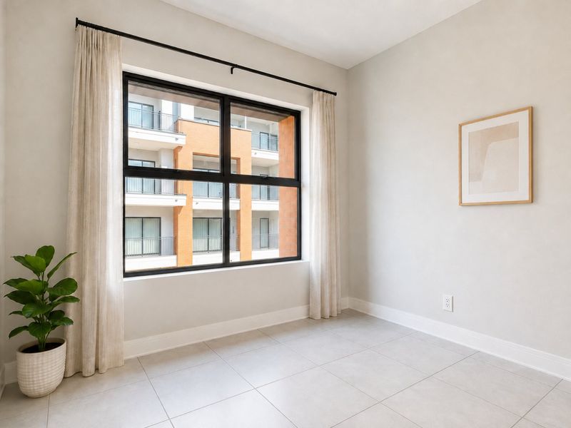 A bright, empty bedroom featuring neutral tiled flooring, clean walls, and a large window with modern black framing, offering a view of the complex.