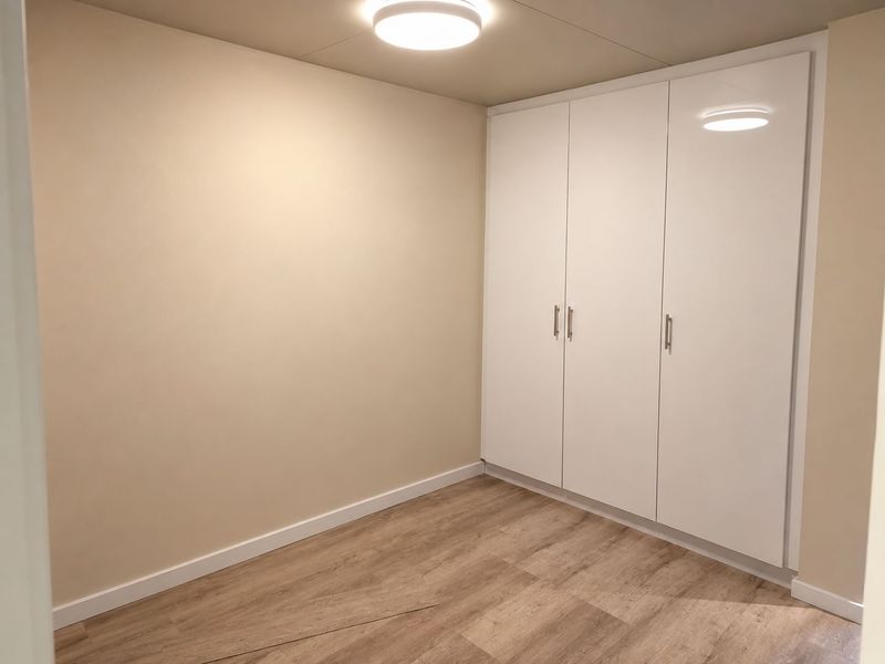 A clean, empty bedroom featuring modern white built-in wardrobes, light-toned laminate flooring, and neutral wall finishes.