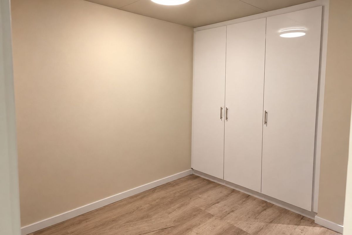 A clean, empty bedroom featuring modern white built-in wardrobes, light-toned laminate flooring, and neutral wall finishes.