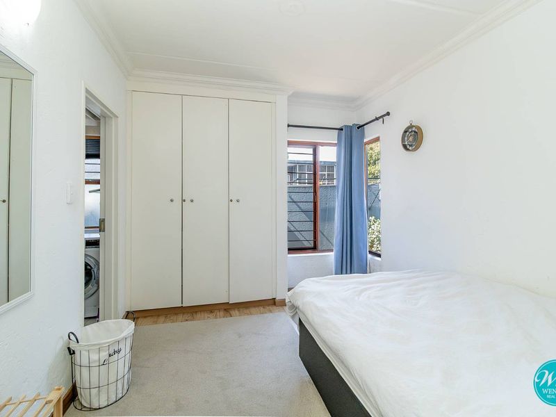 A bright bedroom featuring built-in cupboards, timber window frames, and classic cornicing, with an adjacent laundry area visible through the doorway.