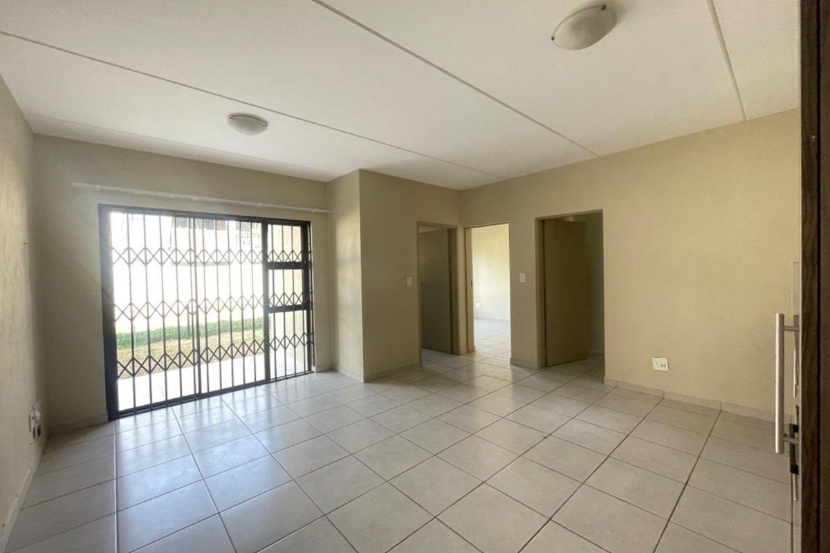An empty, open-plan living area featuring neutral tiled flooring, a sliding door with a security gate, and access to adjacent rooms.