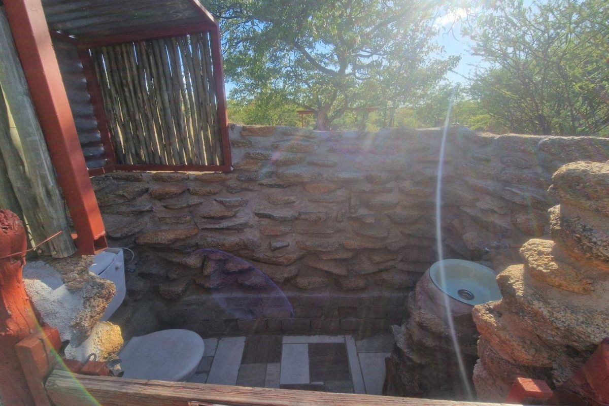 An outdoor rustic bathroom featuring natural stone walling, a built-in stone basin, and a toilet, set within a natural lodge environment.