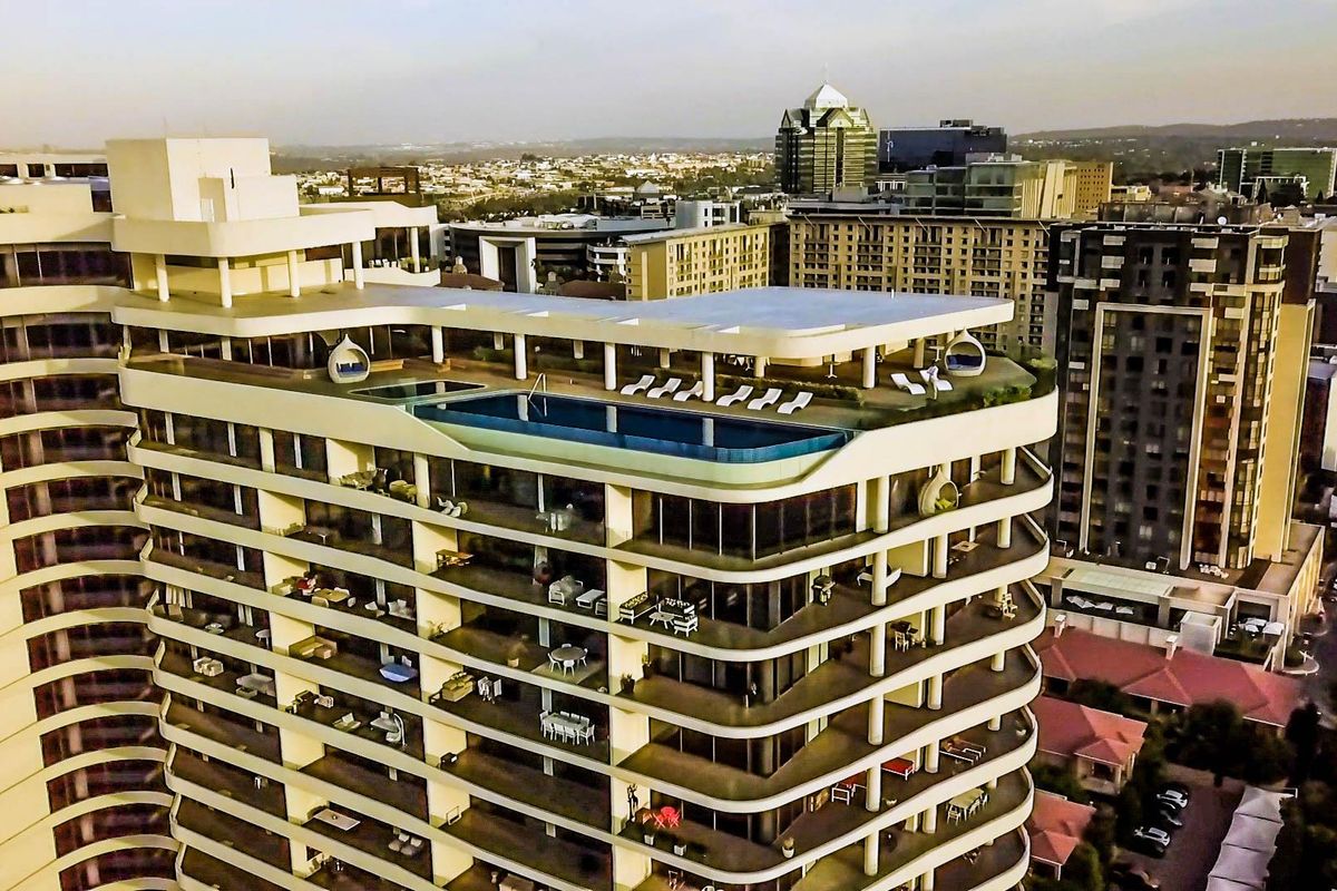 Aerial view of a modern high-rise penthouse building featuring a rooftop swimming pool and expansive wrap-around balconies with city skyline views.