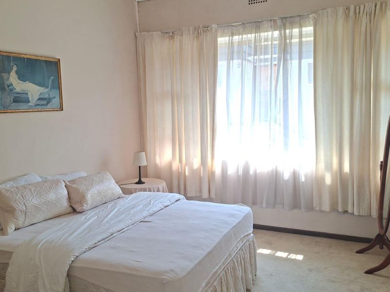 A well-lit bedroom featuring neutral wall tones, carpeted flooring, and a large window with curtain fixtures.