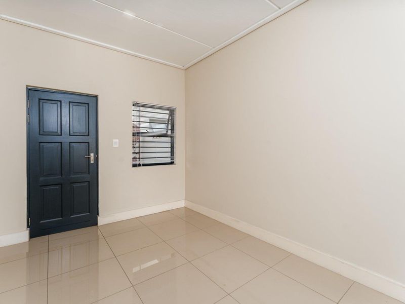 A clean, empty bedroom featuring neutral wall paint, polished floor tiles, and a dark-toned panel door with a small window fitted with venetian blinds.