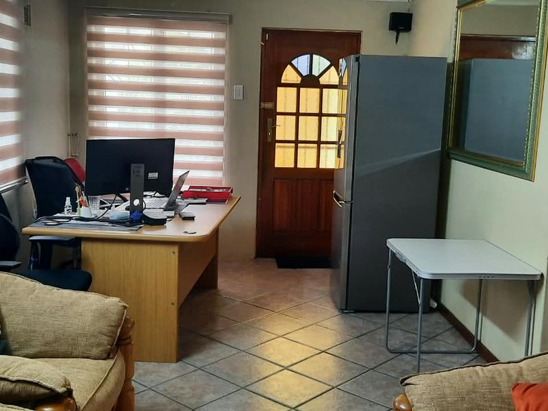 A multi-functional living area featuring tiled flooring, a timber entrance door, and roller blinds, currently utilized as a combined lounge and home office space.