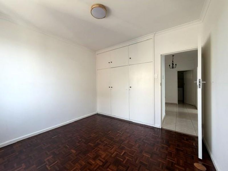 A bright, empty bedroom featuring classic parquet flooring and ample built-in cupboard storage, leading into a tiled hallway.