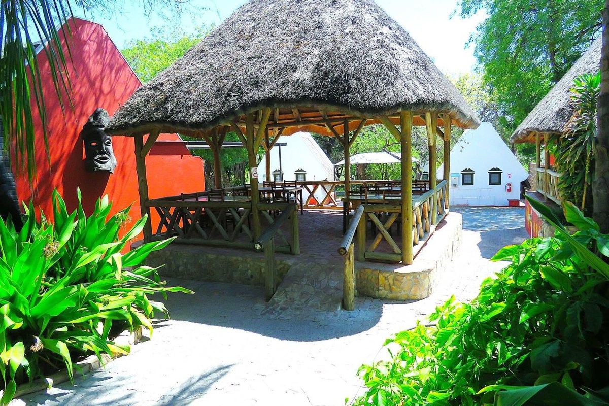 A rustic outdoor gazebo featuring a traditional thatched roof, timber framing, and stone base, set within a landscaped garden area.