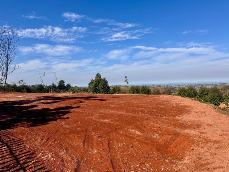 A vacant residential plot featuring cleared red earth and expansive distant landscape views under a clear blue sky.