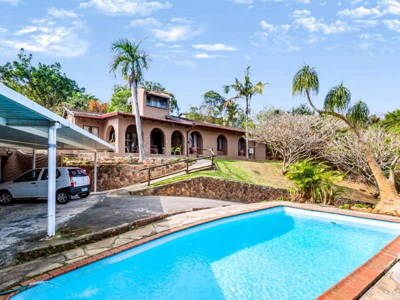 Rear exterior view featuring a swimming pool, stone retaining walls, a covered carport, and a house with an arched veranda.