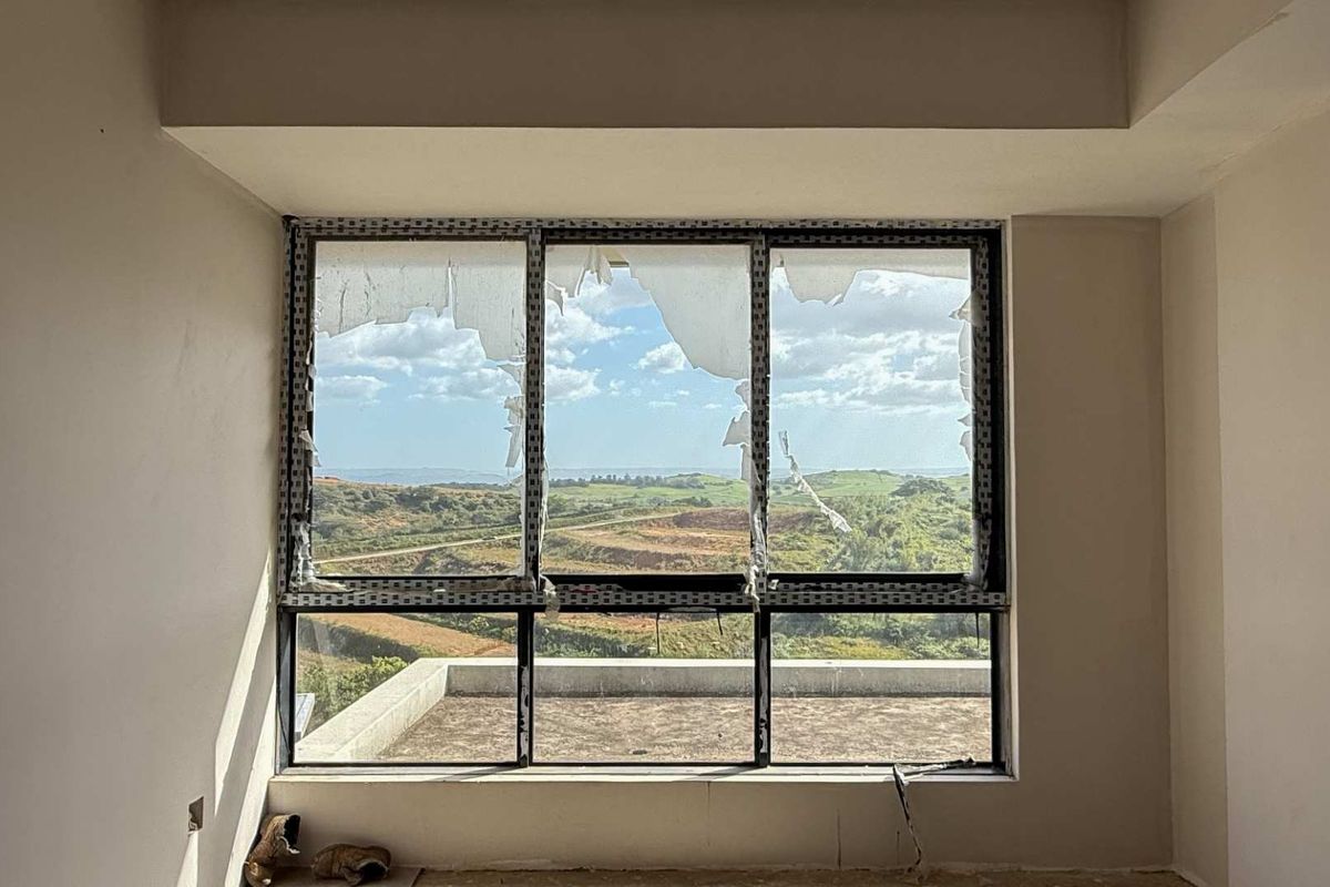 An interior room featuring a large window with significant glass damage, offering expansive views of the surrounding landscape.