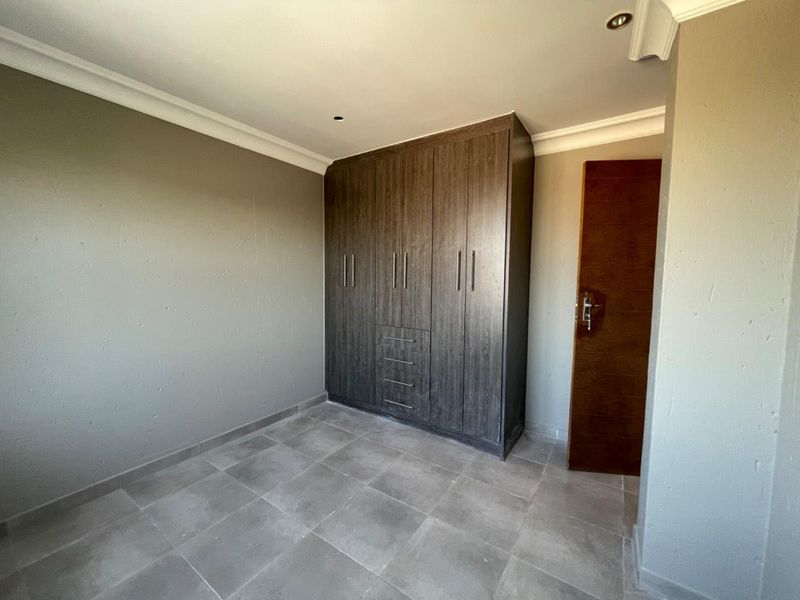 A clean, empty bedroom featuring modern dark wood built-in cupboards, neutral grey tiled flooring, and recessed ceiling lighting.