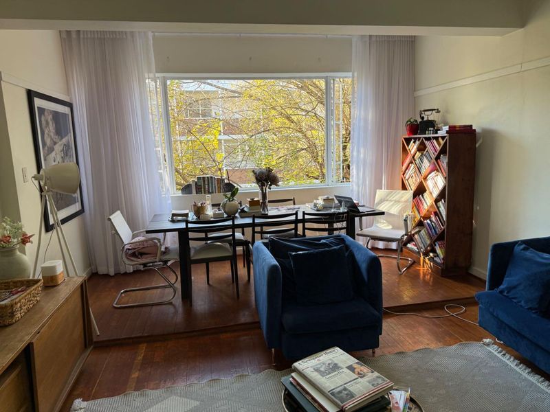 A bright living room featuring original hardwood flooring, a raised dining platform, and a large picture window overlooking mature trees.