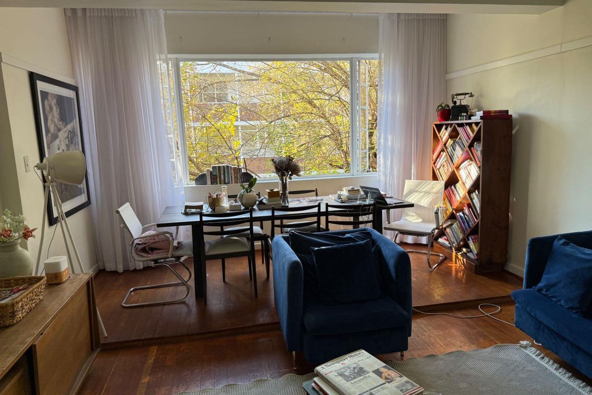 A bright living room featuring original hardwood flooring, a raised dining platform, and a large picture window overlooking mature trees.