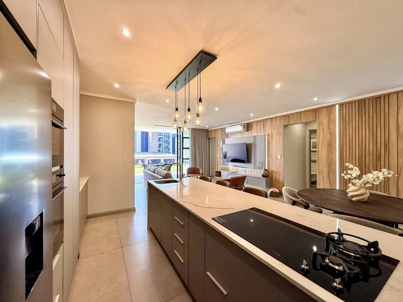 Modern open-plan kitchen and living area featuring a stone-topped island, gas hob, integrated appliances, and elegant timber wall paneling with recessed lighting.