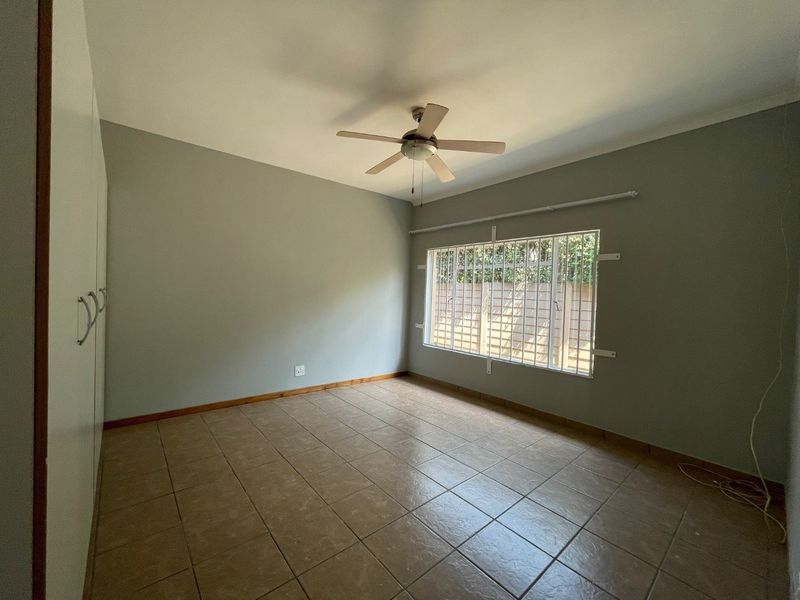 A spacious, empty bedroom featuring tiled flooring, built-in cupboards, and a ceiling fan, with security bars installed on the window.