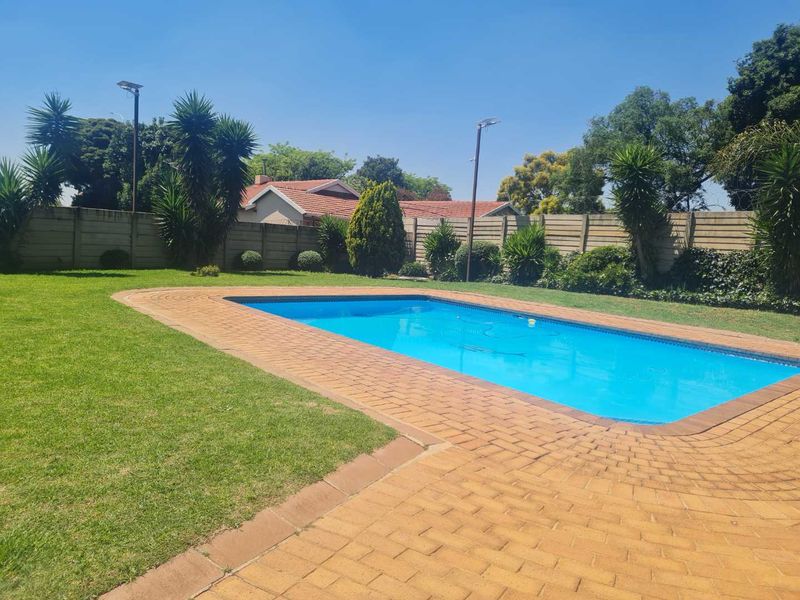 A well-maintained outdoor pool area featuring a clear swimming pool, brick paved surround, and a manicured lawn enclosed by a perimeter wall.