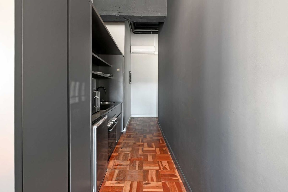 A compact kitchenette featuring dark cabinetry, integrated appliances, and parquet flooring, set within a modern apartment space with exposed industrial-style ceiling details.