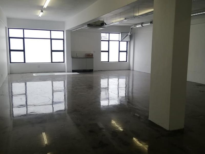 Open-plan office space featuring polished concrete flooring, exposed ceiling services, large windows for natural light, and a small kitchenette area.