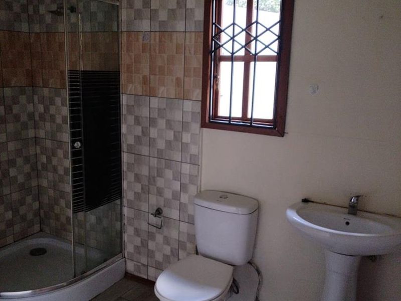 A functional bathroom featuring a corner shower enclosure, pedestal basin, and toilet, with tiled wall accents and a window fitted with a security gate.