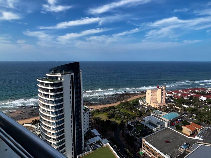 Elevated panoramic coastal view overlooking the ocean, beach, and surrounding urban landscape from a high-rise balcony.