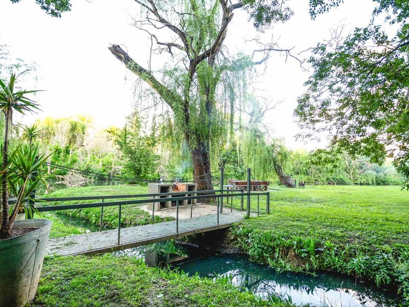 A scenic garden area featuring a small pedestrian bridge over a stream, a built-in braai, and mature trees in a tranquil outdoor setting.