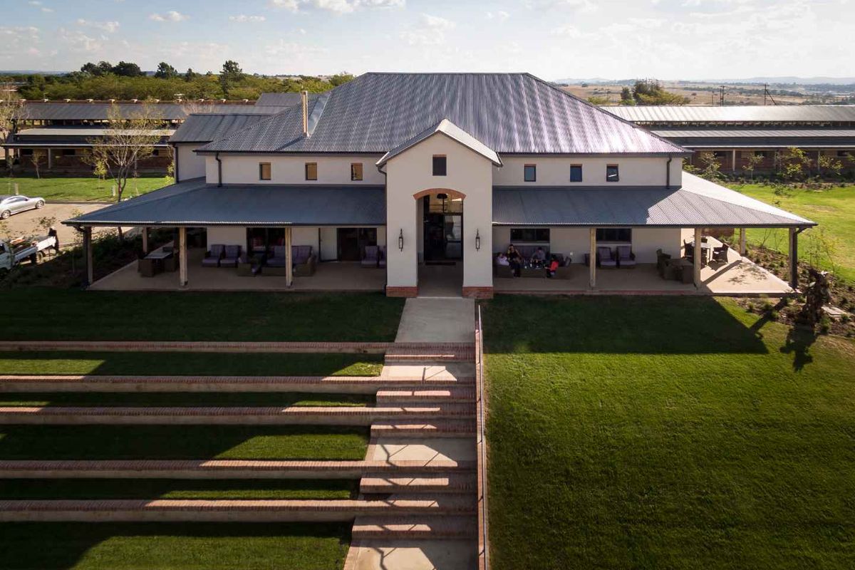 An aerial view of a large, well-maintained farmhouse featuring a covered wrap-around veranda, metal roof, and landscaped terraced lawn with brick steps.