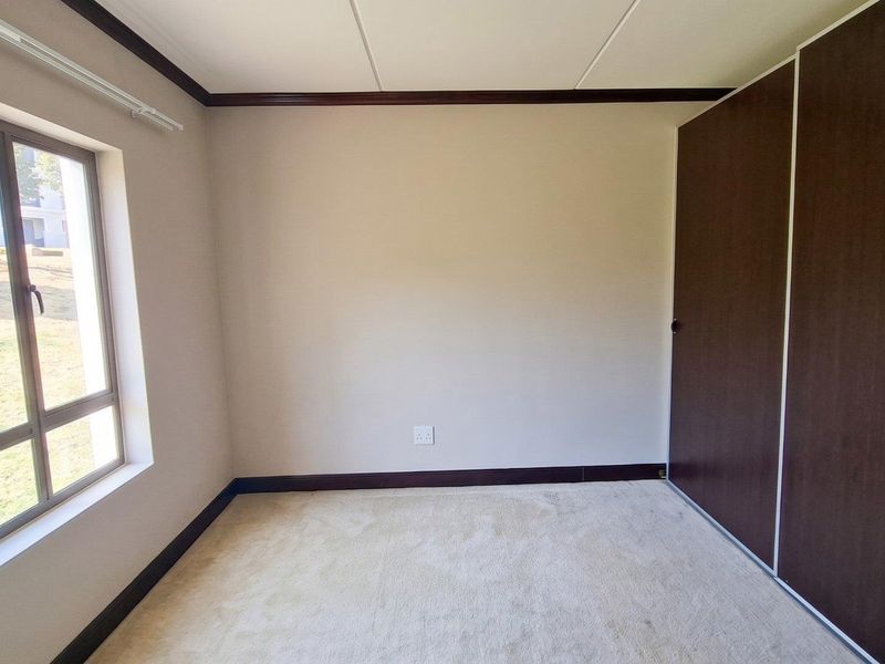 A bright, empty bedroom featuring neutral carpeting, dark wood-finish built-in cupboards, and a large window overlooking the exterior grounds.