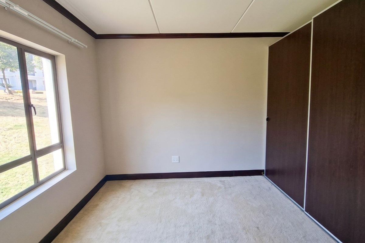 A bright, empty bedroom featuring neutral carpeting, dark wood-finish built-in cupboards, and a large window overlooking the exterior grounds.